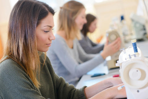 A woman practices sewing on a machine during an adult sewing class, while other students work on their projects in the background.